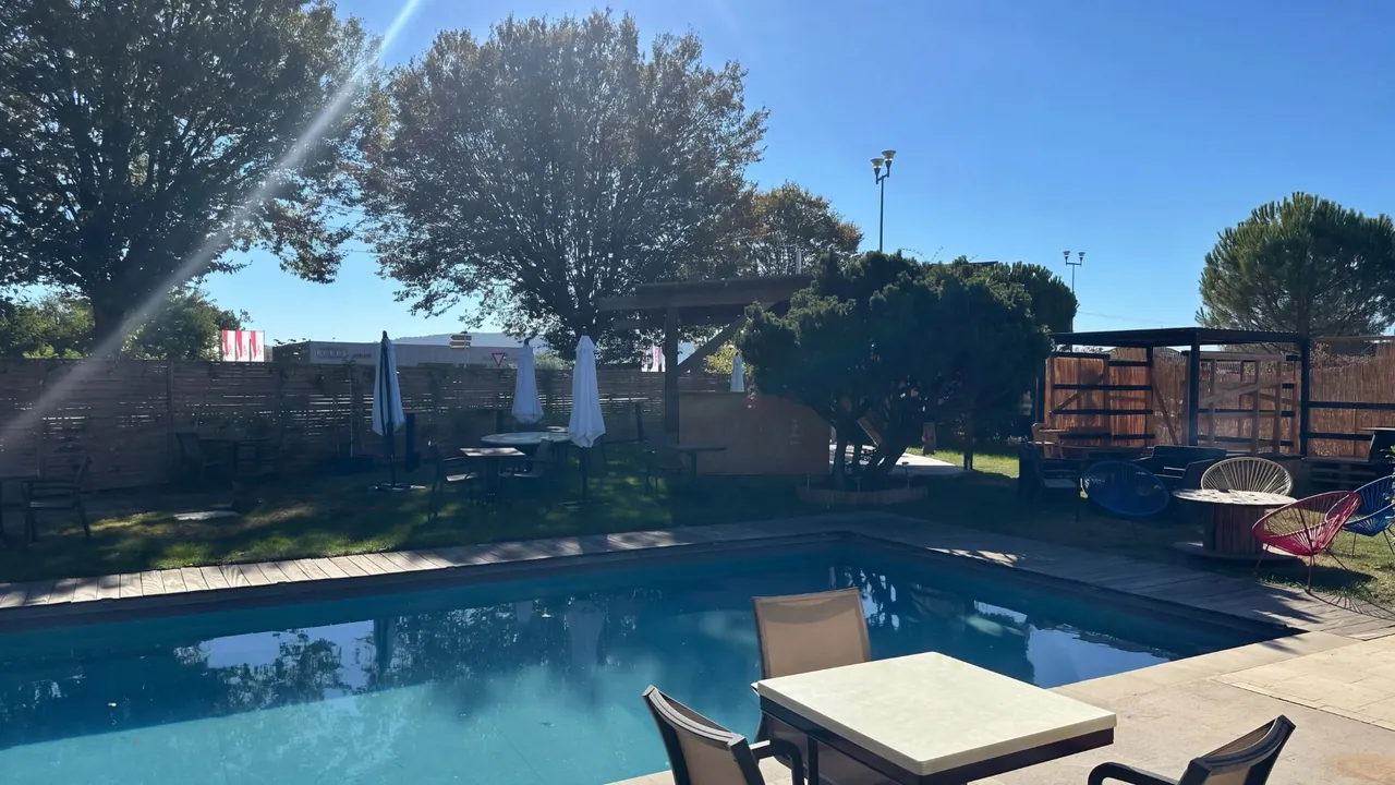 Le bassin en plein soleil, une table au bord de l’eau, les parasols, le petit bar en bois et le coin lounge sous les arbres